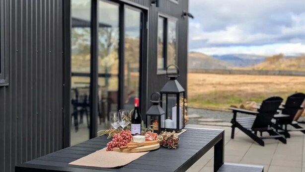 Una mesa dispuesta con botella de vino, copas, uvas, queso y flores en un patio con asientos al aire libre y vistas al paisaje.