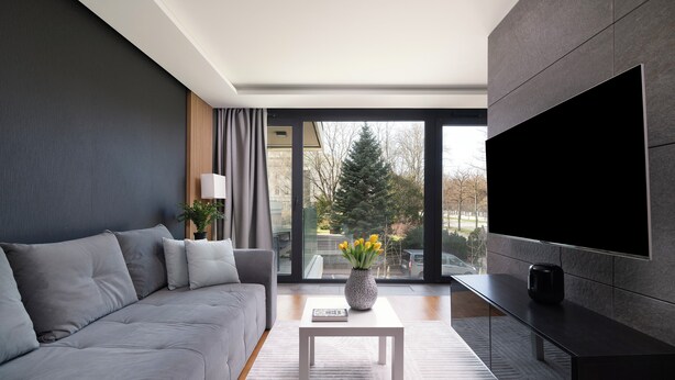 A modern living room with a grey sofa, a white coffee table with a vase of flowers, and a large flat-screen TV mounted on the wall. - EASY RENT Apartments - AMBER 10 (Lublin)
