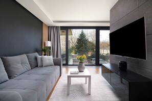 A modern living room with a grey sofa, a white coffee table with a vase of flowers, and a large flat-screen TV mounted on the wall. - EASY RENT Apartments - AMBER 10 (Lublin)