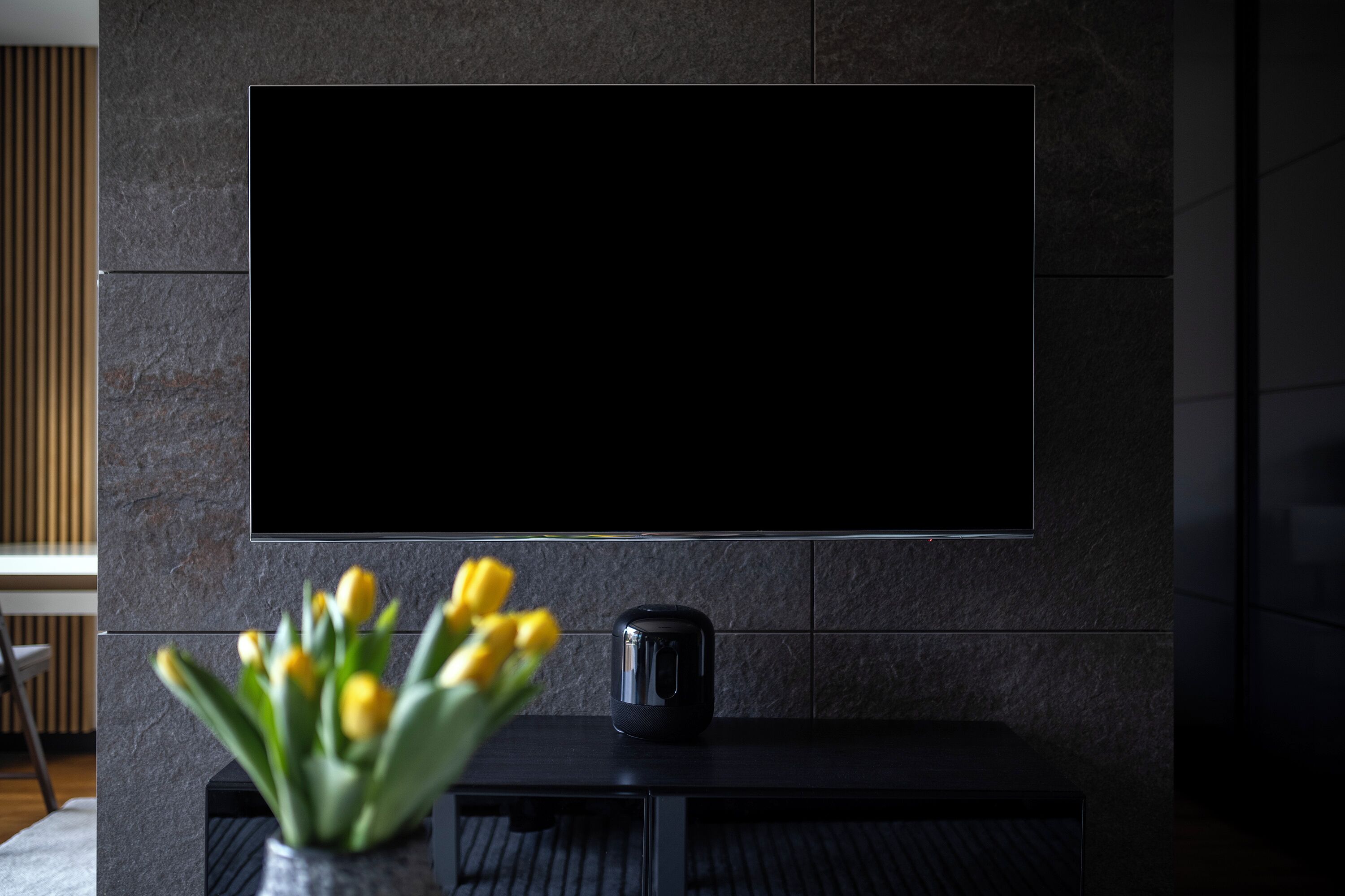 Un soggiorno moderno con TV a schermo piatto montata su una parete strutturata, un tavolino da caffè scuro e un vaso con fiori gialli.