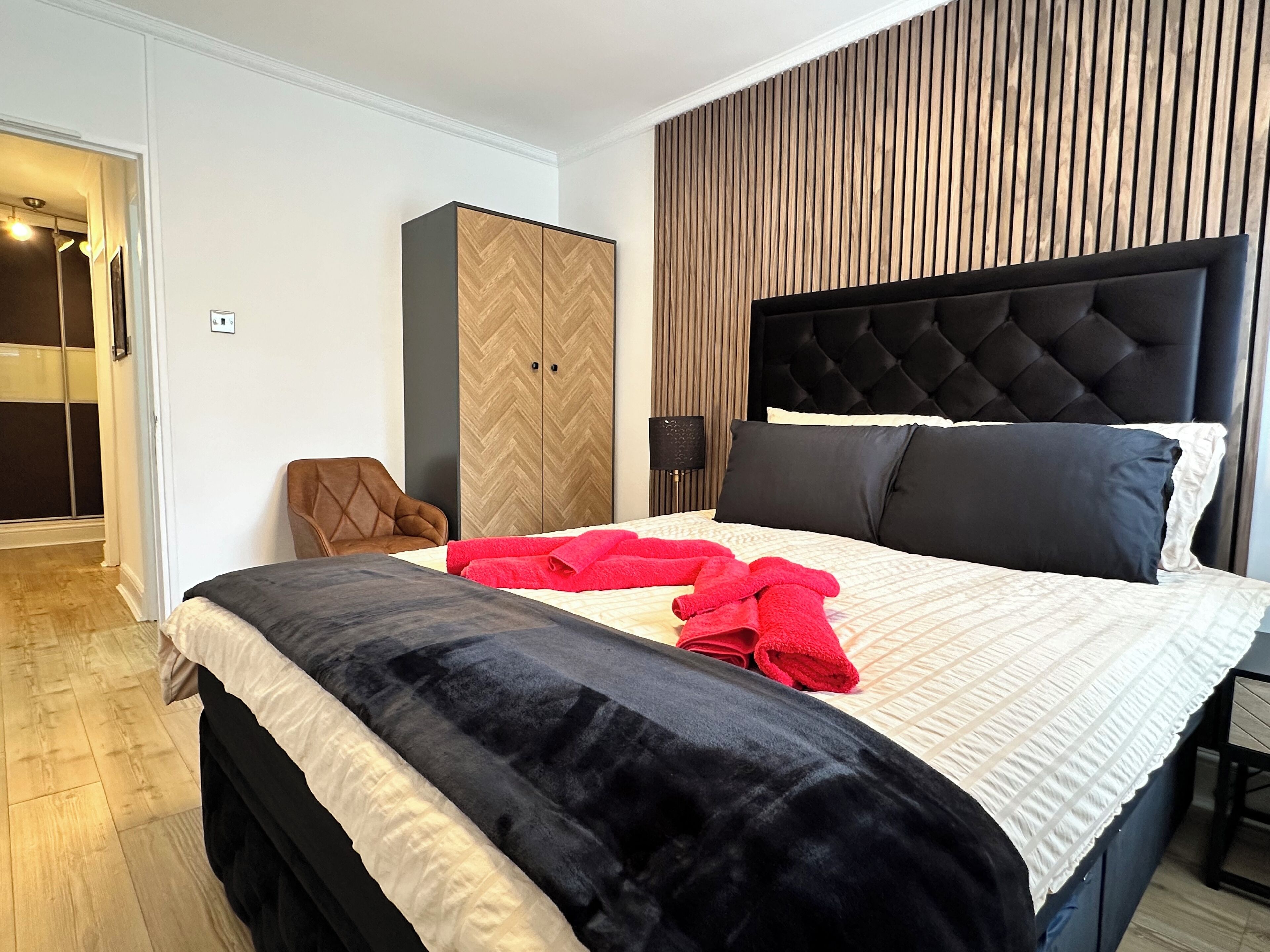 A modern hotel room with a large bed, a wooden wardrobe, a brown leather chair, and a headboard with a tufted design.