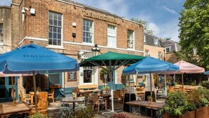 An outdoor seating area with tables and chairs under umbrellas, a brick building with 'THE WINDMILL' sign, and a clear sky. - Clapham 3-bed Flat, Sleeps 6 With Free Parking (London)
