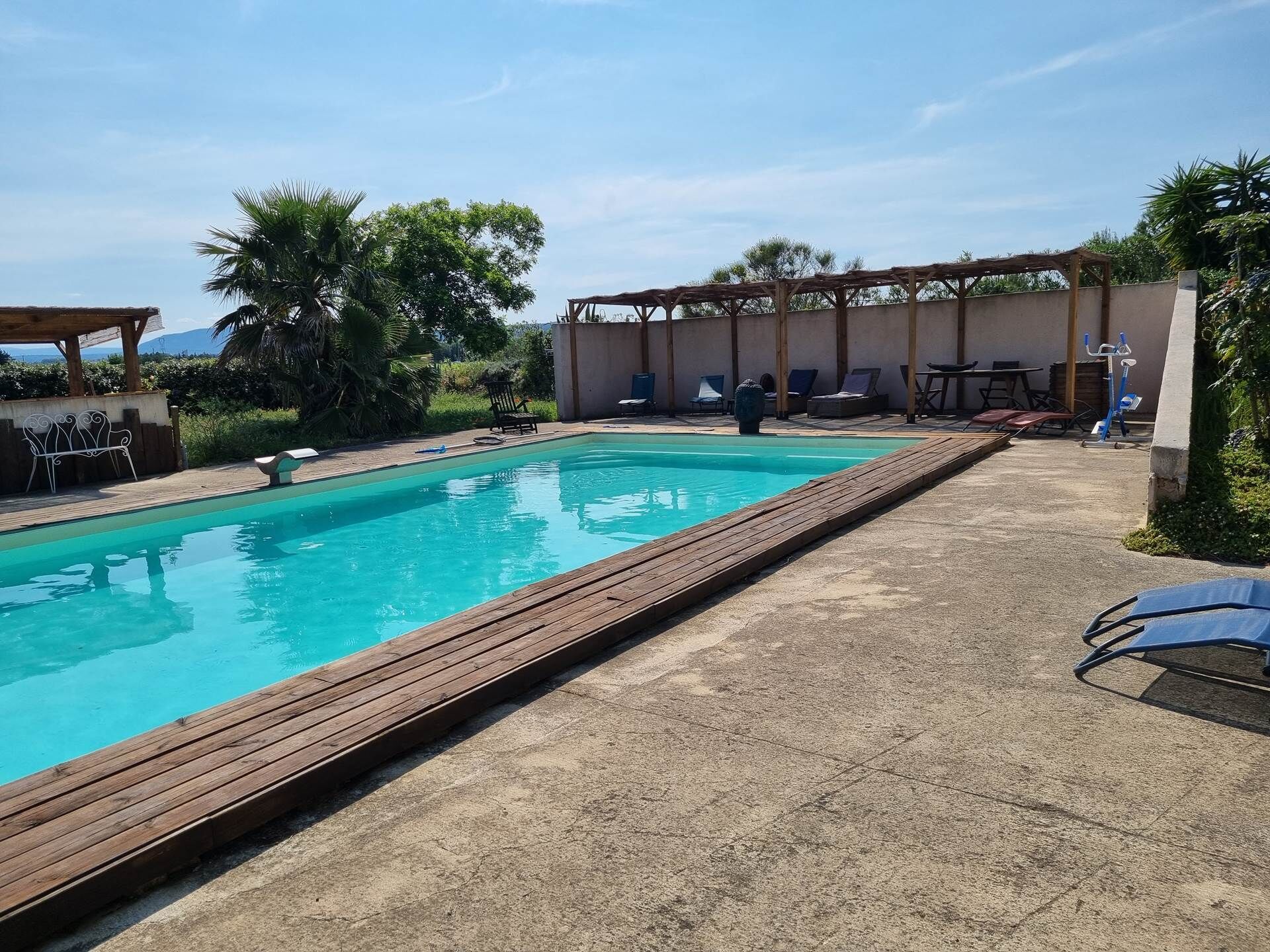 Een zwembad met een houten terras, ligbedden en een overdekt gedeelte met tafels en stoelen.