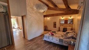 A bedroom with a wooden floor, a bed with patterned bedding, a hanging light fixture, and a bathroom with a mirror and sink. - Large Gîte 2 people (LEZIGNAN CORBIERES)