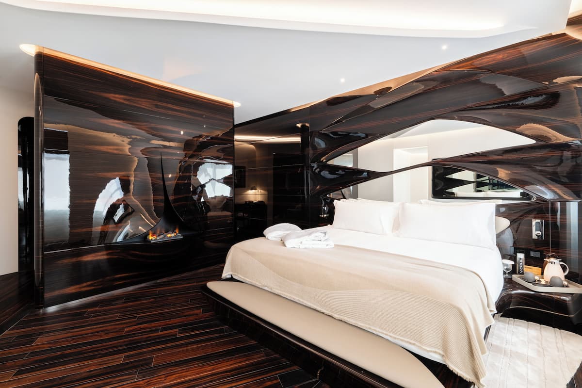 A modern hotel room with a large bed, a bench, a fireplace, and a large mirror.