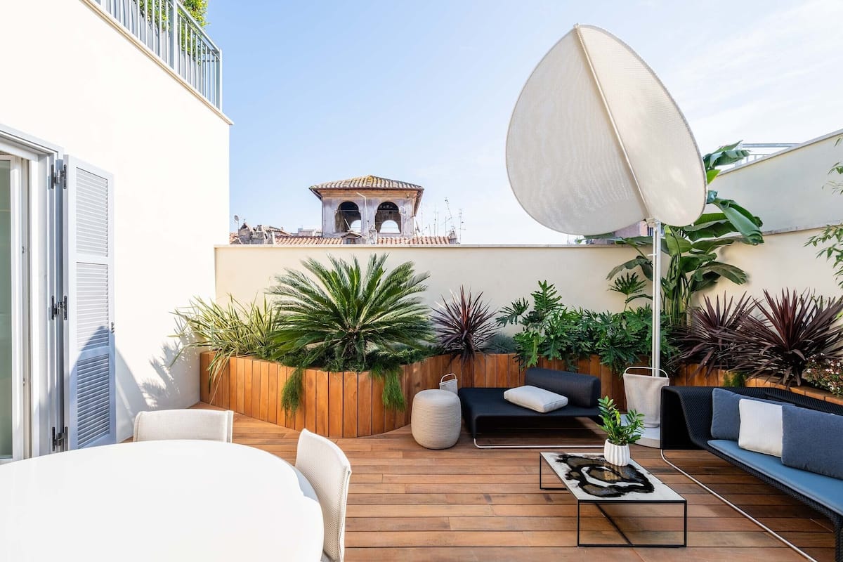 A rooftop patio with wooden flooring, a black sofa, a white table, and various potted plants.