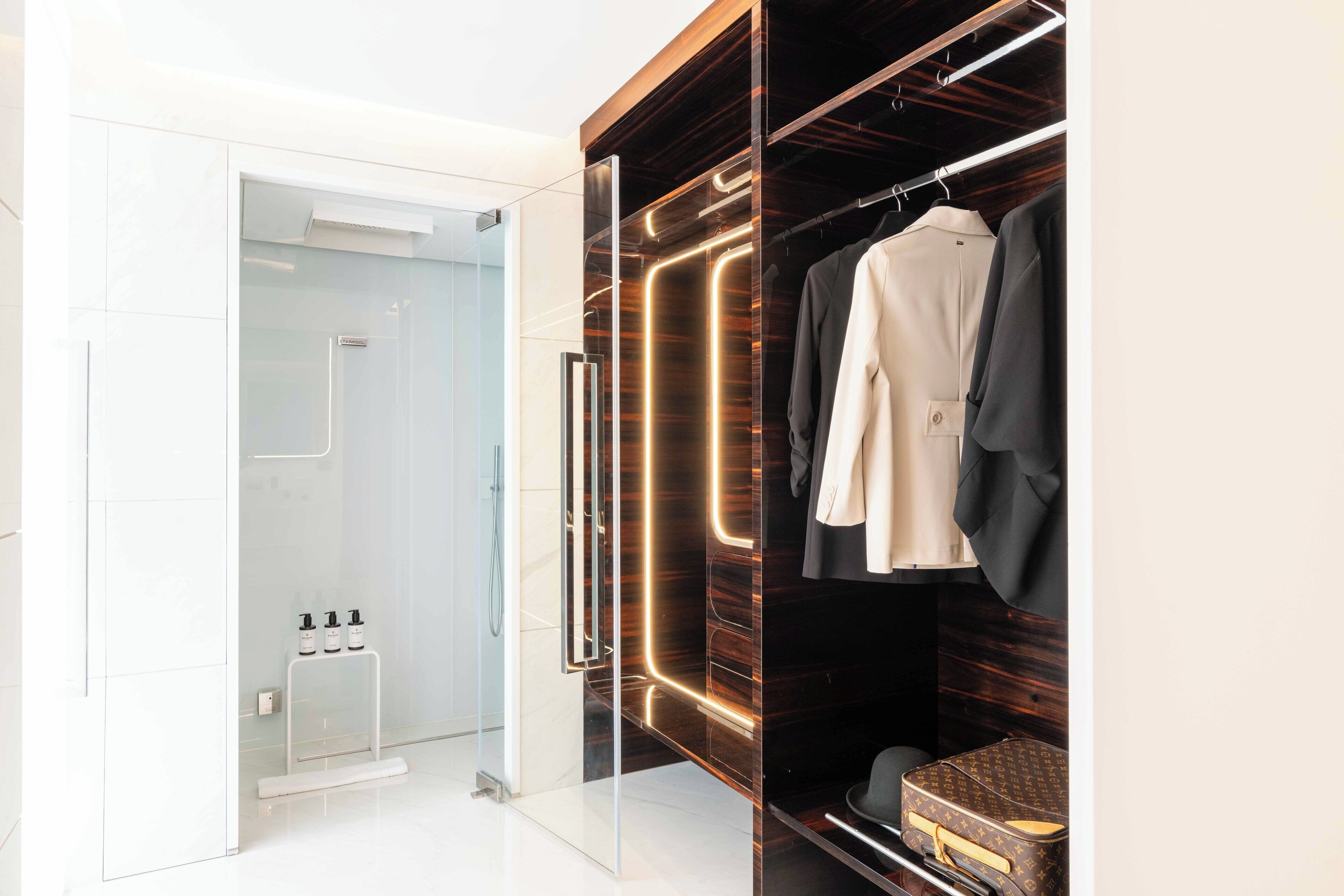 A modern bathroom with a glass shower enclosure, a vanity with a mirror, and a walk-in closet with a wooden finish and illuminated lighting.