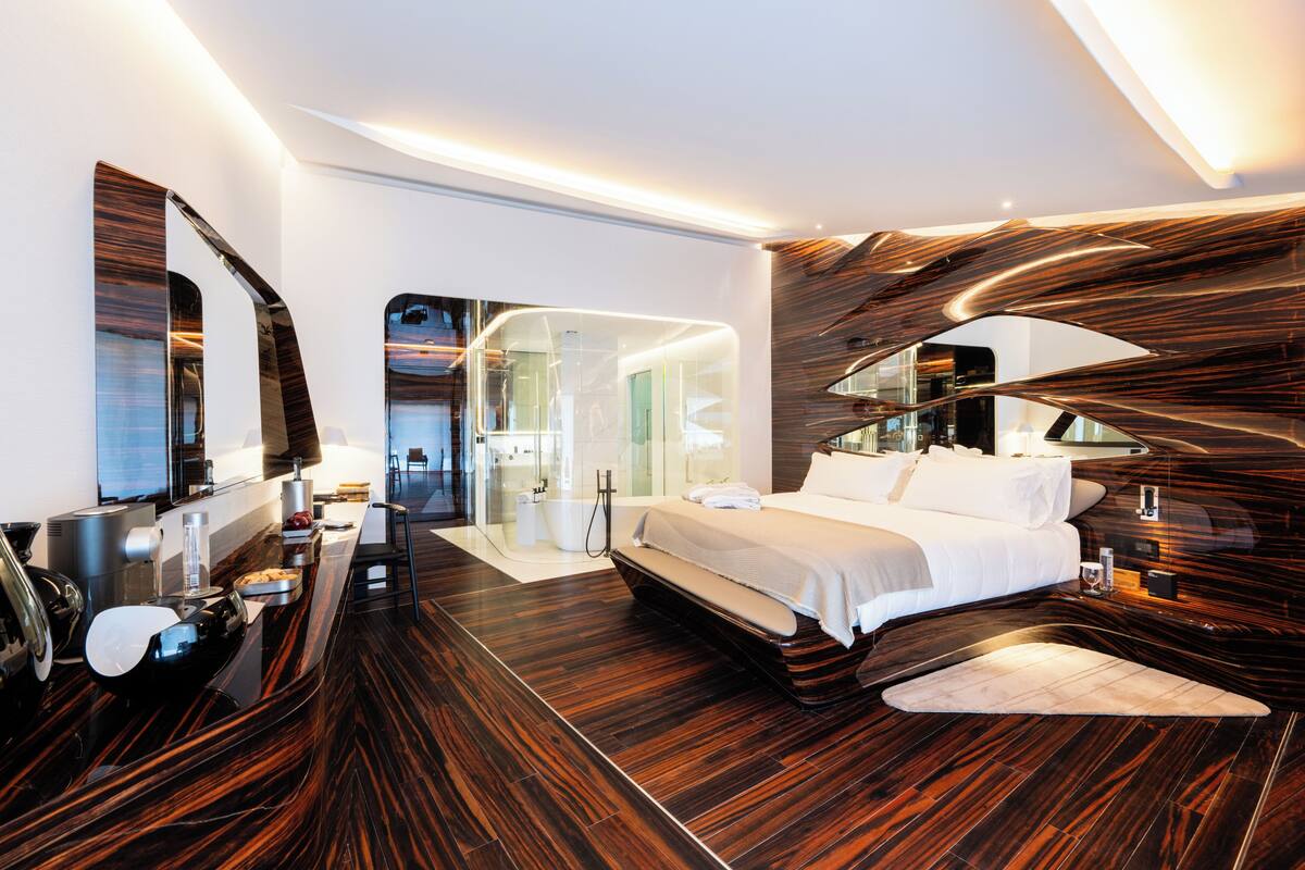 A modern hotel room with a large bed, a desk with a chair, a large mirror, and a bathroom with a glass shower enclosure.