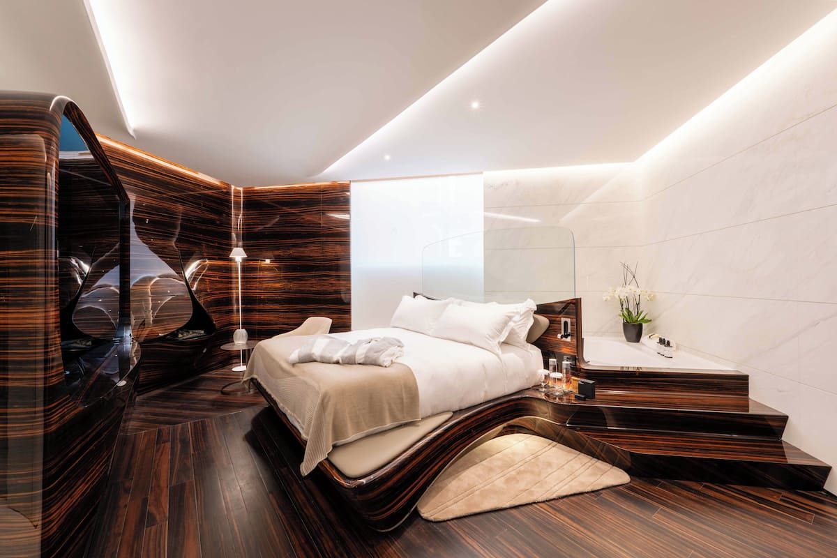 A modern bedroom with a curved bed, a bedside table, a nightstand, a mirror, and a bathroom with a sink and mirror.