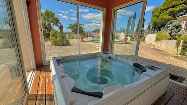 A hot tub in a room with large windows, a wooden deck, and a view of trees and hills. - Groupe 12 Personnes sur 3 Gîtes (LEZIGNAN CORBIERES)