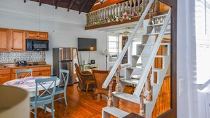 A kitchen with wooden cabinets, a microwave, and a refrigerator. There is a dining area with a table and chairs. A staircase with a railing is visible in the background. - Rose Key Cottage - Private Pool & Near Duval (Key West)