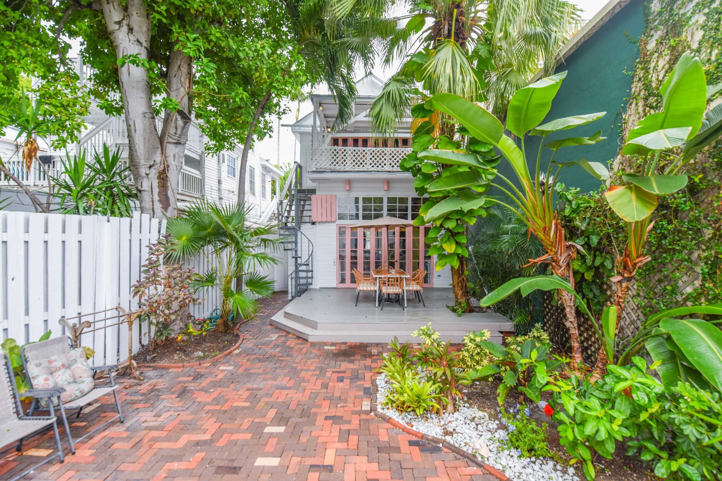 A courtyard with a brick pathway, a white fence, a wooden bench, and a dining area with chairs and a table.