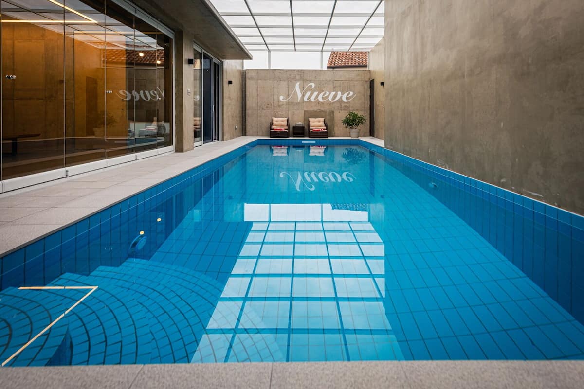 Ein moderner, überdachter Pool mit klaren, blauen Fliesen, einer Betonwand mit der Aufschrift 'Nueve' und einem Glastür-Eingang.
