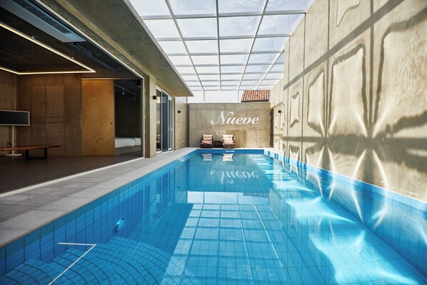An indoor swimming pool with a glass ceiling, a wall with the word 'Nueve' written on it, and a bench with pillows. - Nueve Poolvilla And Resort (Jeju City)