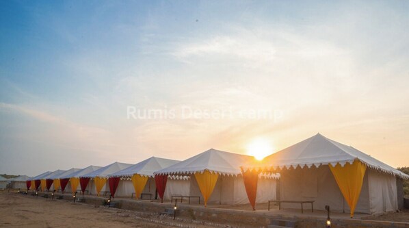 A row of tents with white canopies and red and yellow curtains, set against a desert backdrop with the sun setting. - Rumis Desert Camp (Jaisalmer)