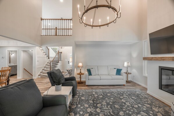 A spacious living room with a sofa, a coffee table, and a chandelier. There is a staircase leading to an upper level.