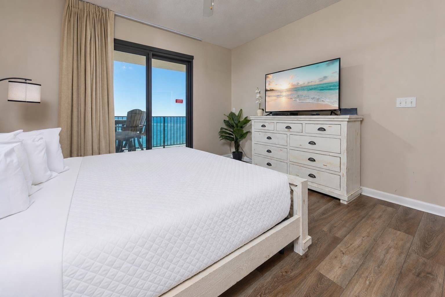 A hotel room with a large bed, a dresser, a television, and a balcony with a view of the ocean.