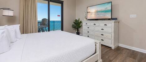 A hotel room with a large bed, a dresser, a television, and a balcony with a view of the ocean.