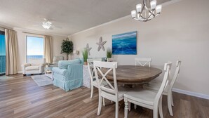 A spacious living room with a large window, a dining area with a round table and white chairs, and a ceiling fan.