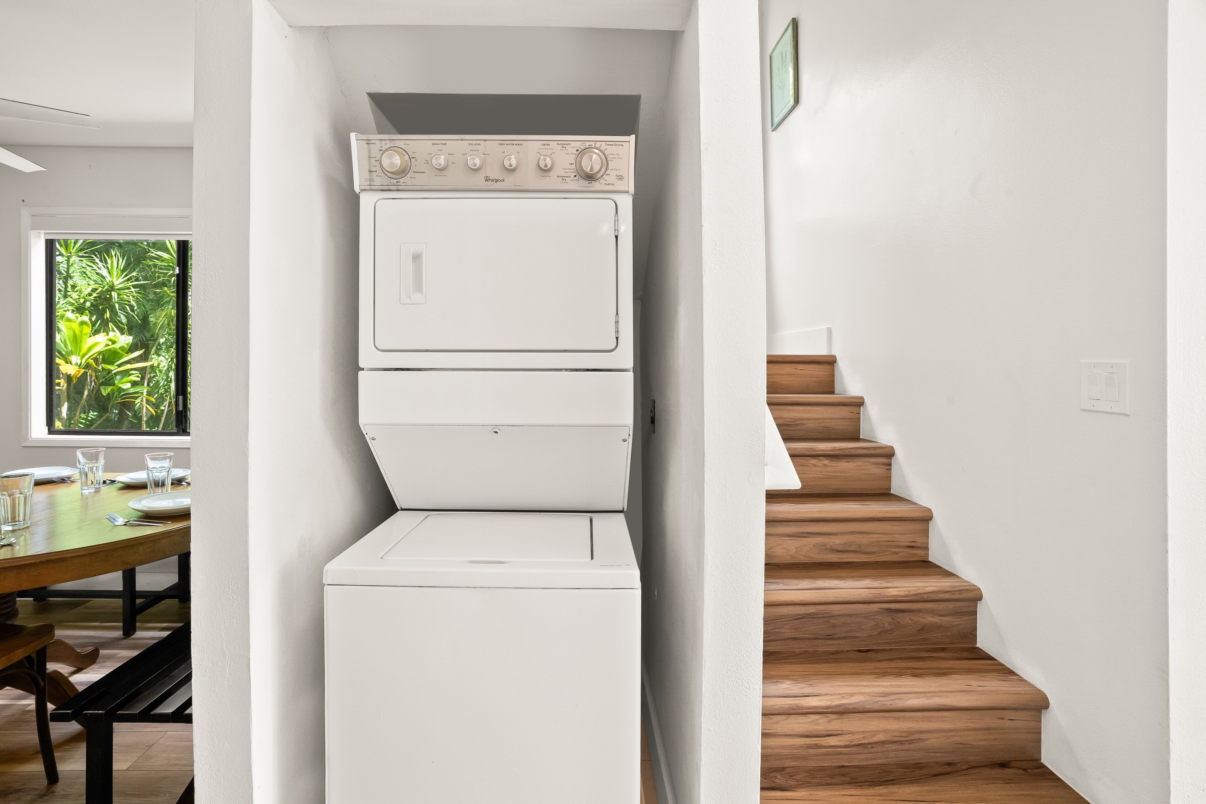 Une petite buanderie avec des lave-linge et des sèche-linge superposés, un escalier en bois et un coin repas offrant une vue sur la verdure.