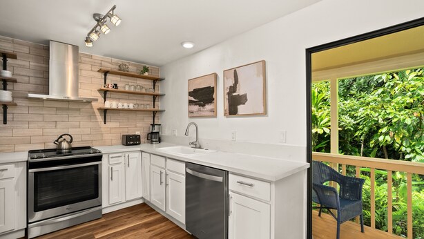 A modern kitchen with white cabinets, a stainless steel stove, and a dishwasher. There are two framed artworks on the wall and a view of lush greenery through a balcony door.