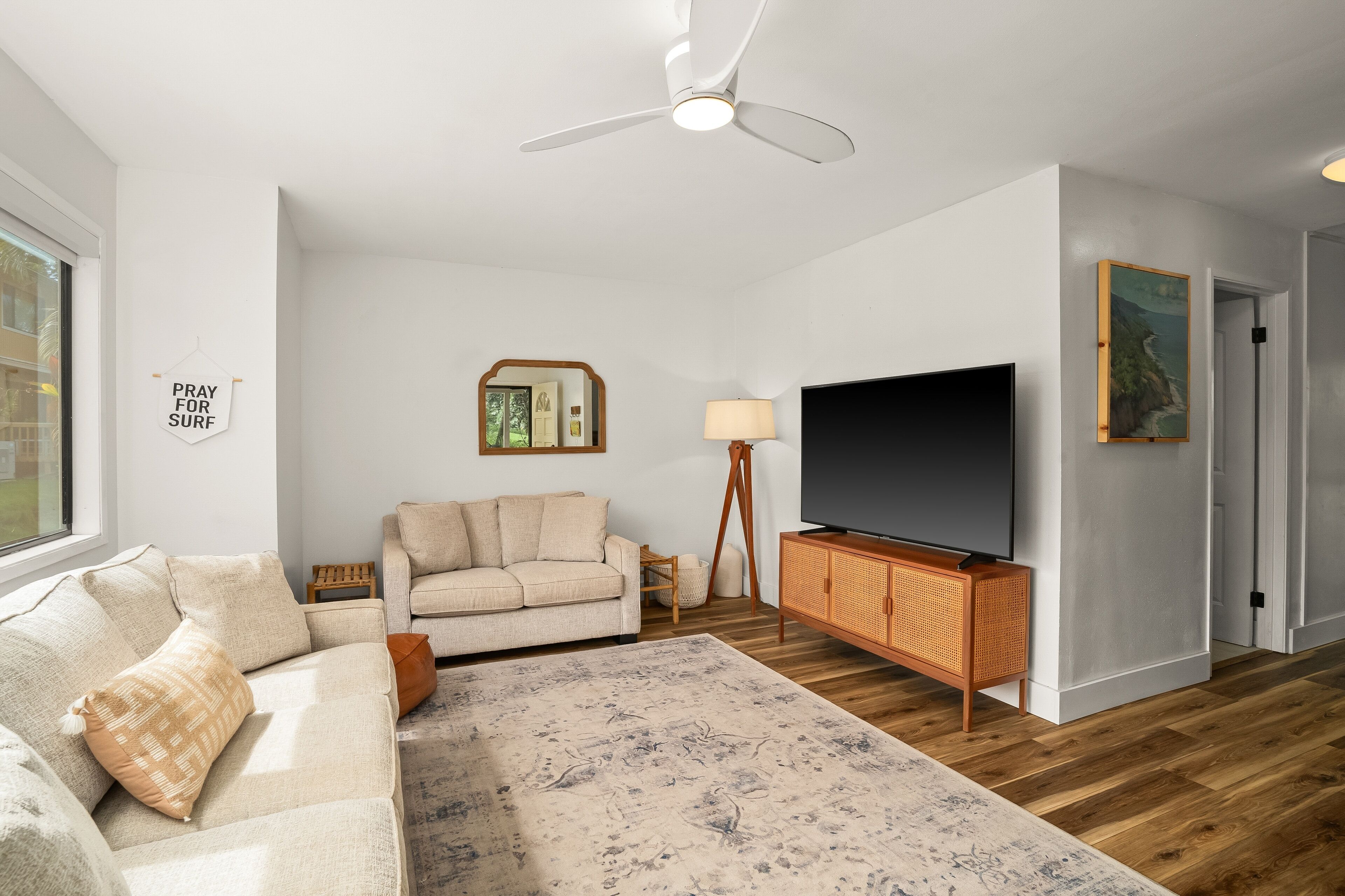 A living room with a beige sofa, a flat-screen TV, a wooden cabinet, a standing lamp, and a ceiling fan.