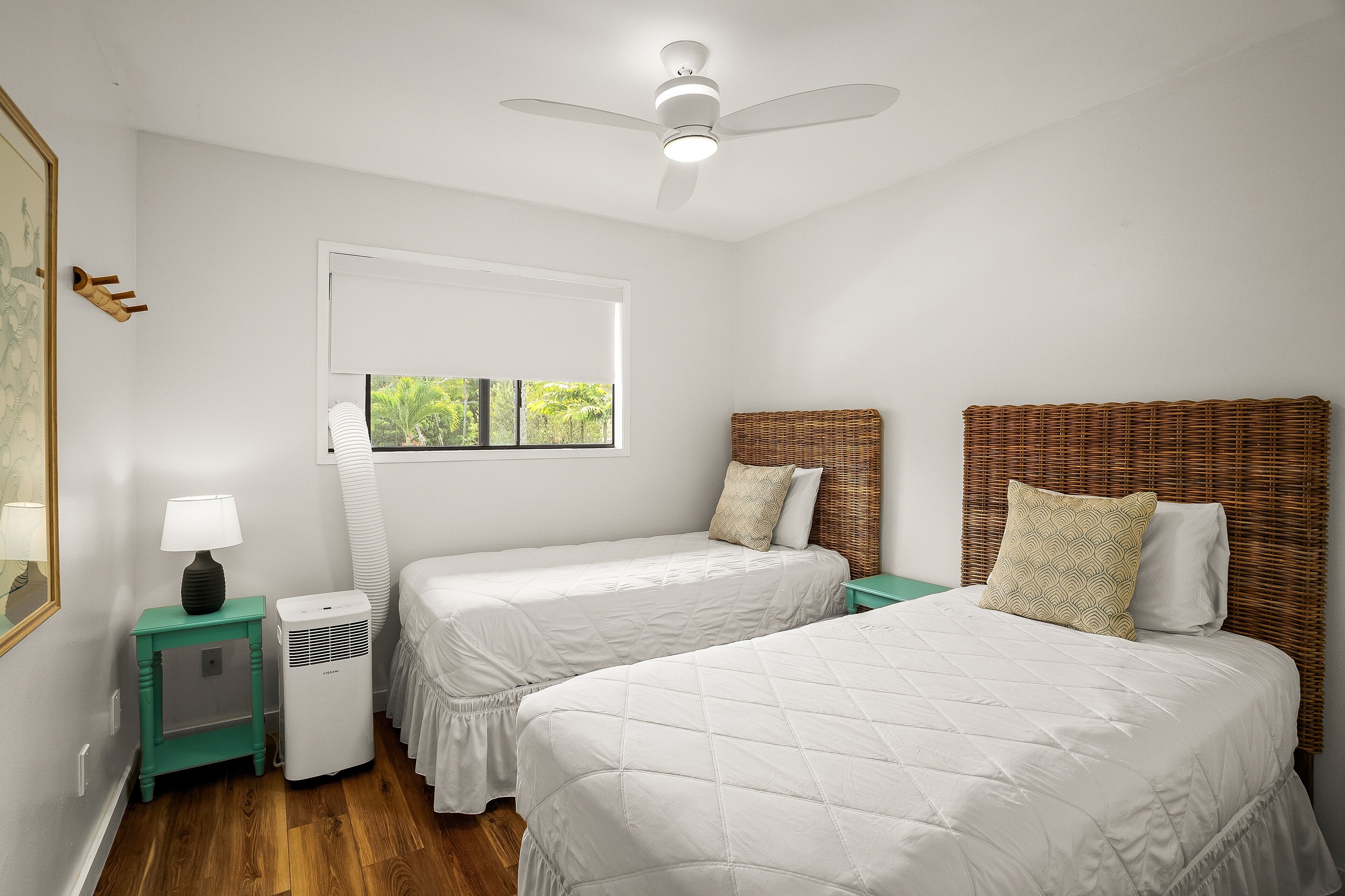 Ein Zimmer mit zwei Betten, einem Deckenventilator, einem Fenster mit Jalousien, einem kleinen Tisch mit Lampe und einem fest montierten Heizkörper.