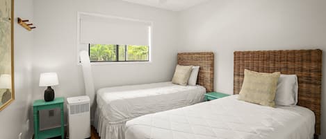 Ein Zimmer mit zwei Betten, einem Deckenventilator, einem Fenster mit Jalousien, einem kleinen Tisch mit Lampe und einem fest montierten Heizkörper.