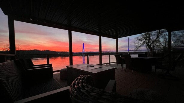 An outdoor seating area with a view of a sunset over a body of water, featuring a table and chairs.