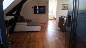 A room with a white sofa, a television mounted on the wall, a kitchen area with cabinets, and a wooden floor. - Zoetermeer City Centre Loft Apartment (Zoetermeer)