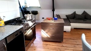 A compact kitchen with a stove, oven, and countertop. There is a wooden cabinet with drawers and a small table with a box and a red item on it. A grey sofa is in the background. - Zoetermeer City Centre Loft Apartment (Zoetermeer)