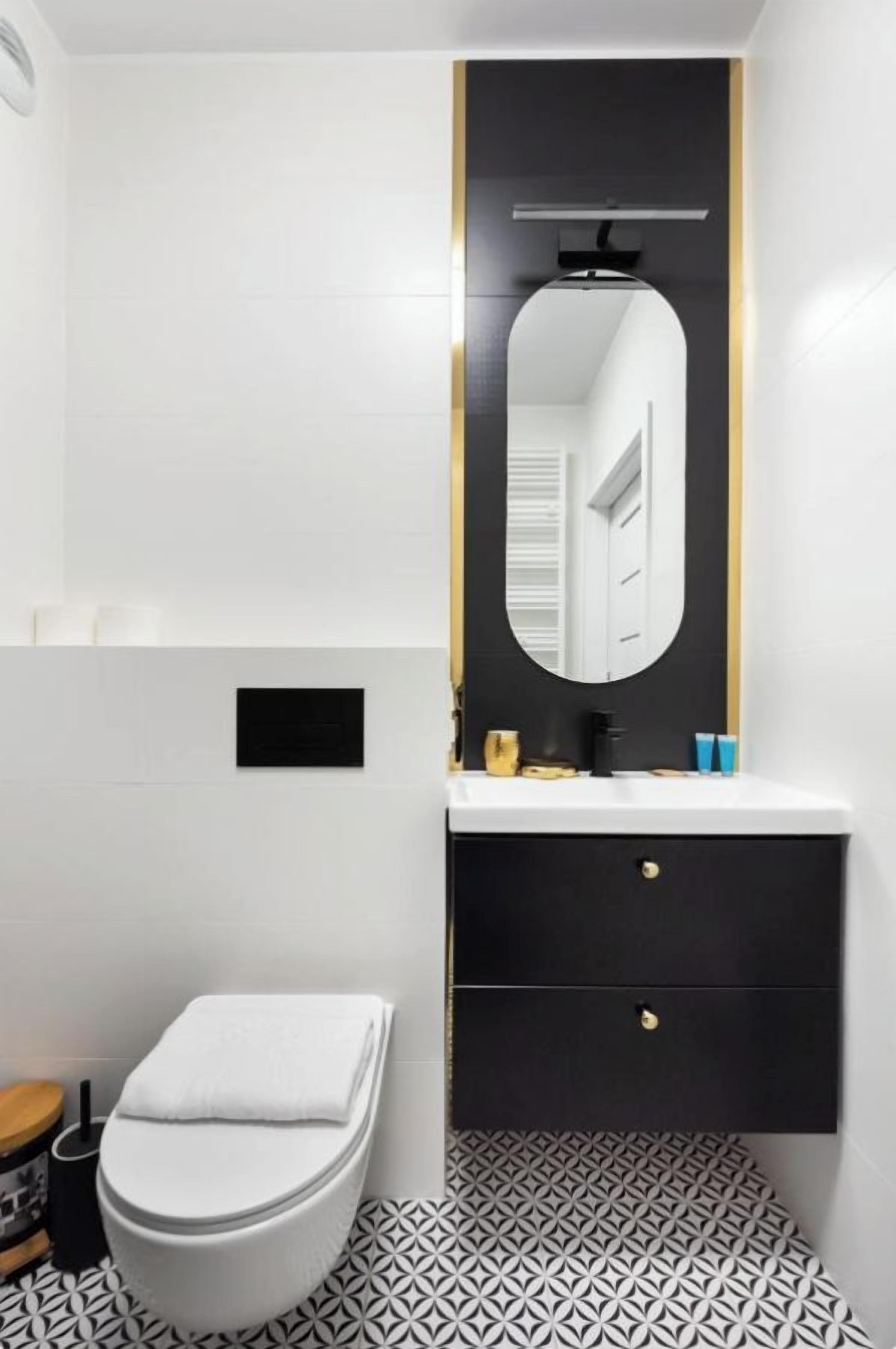 Un baño moderno con inodoro blanco, tocador negro con espejo y baldosas de suelo con diseño.