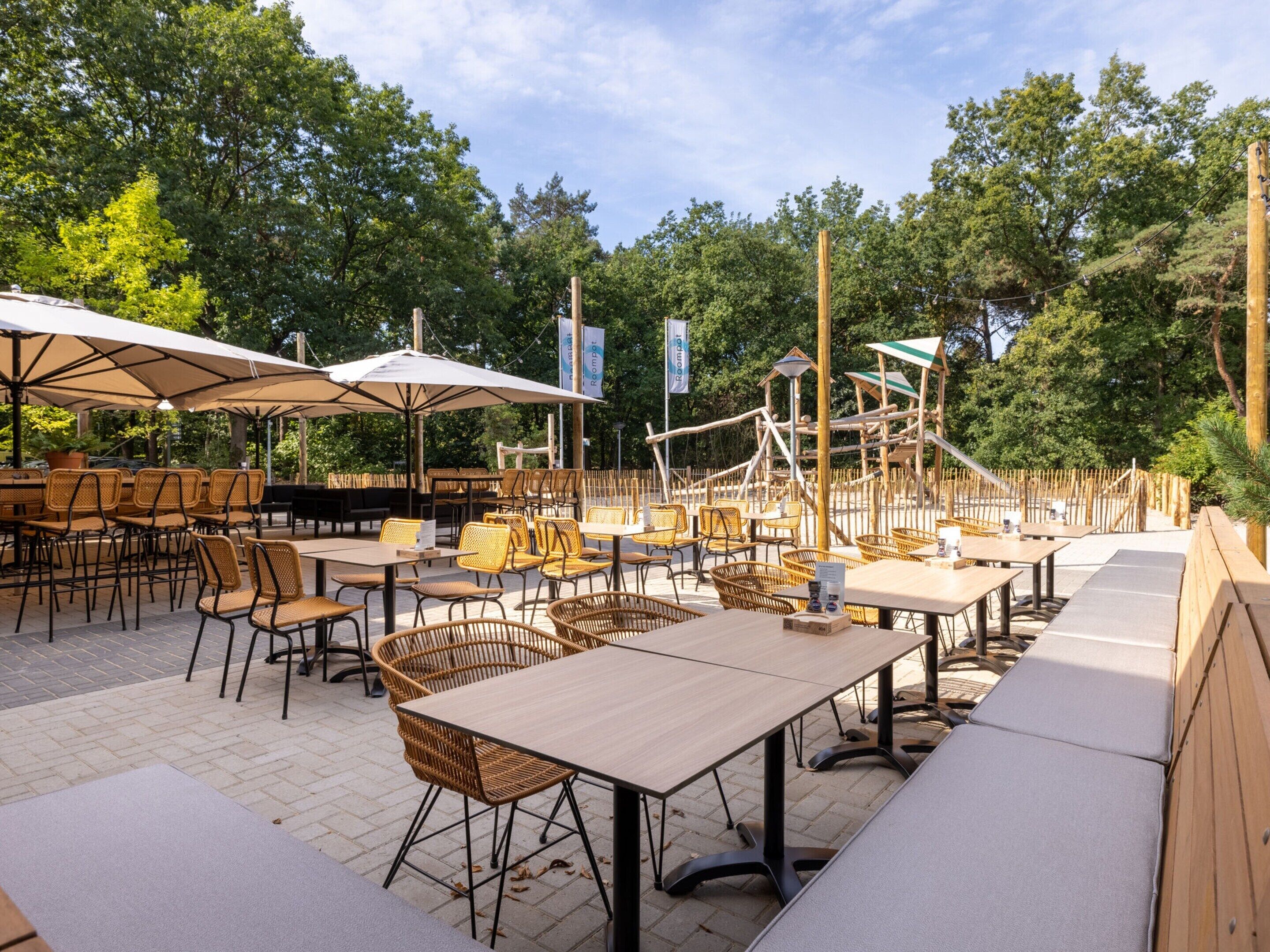 An outdoor dining area with wicker chairs and tables, surrounded by trees and a wooden fence.