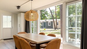 A dining area with a wooden table and chairs, a woven pendant light, and large windows offering a view of the outdoors. - Chalet in De Peel Near Forest (Vlierden)