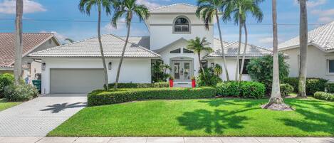 A residential house with a white exterior, a garage, and a well-maintained lawn with palm trees.