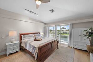 A bedroom with a wooden bed, white nightstands, a ceiling fan, and a view of palm trees through large windows. - S Exposure Waterfront Home | Near Beach/Food/Fun (Marco Island)