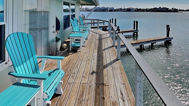 A wooden deck with turquoise Adirondack chairs, a railing, and a view of the water and distant buildings.