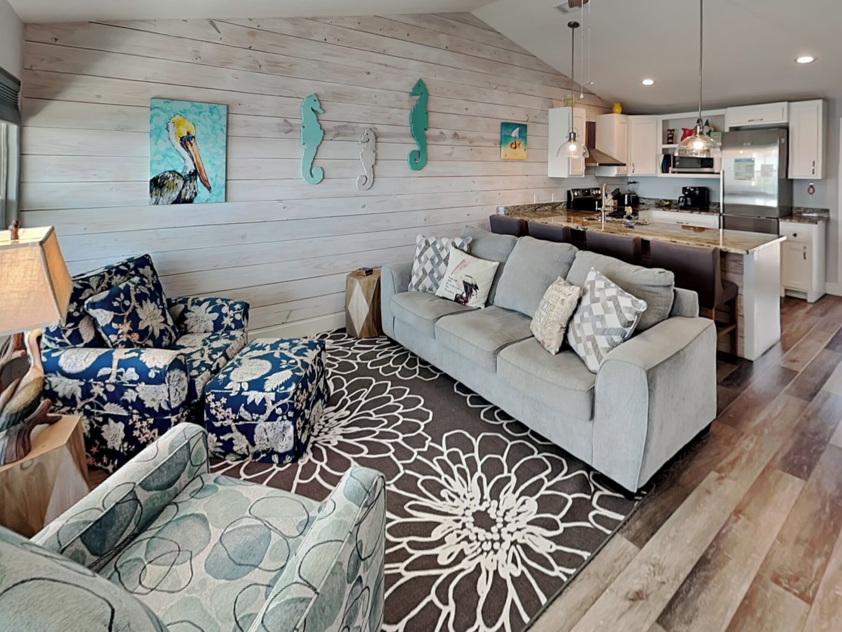 Een moderne woonkamer met een grijze bank, gestreepe fauteuils en een decoratief vloerkleed. Achtergrond toont een keuken met witte kasten en roestvrijstalen apparatuur.
