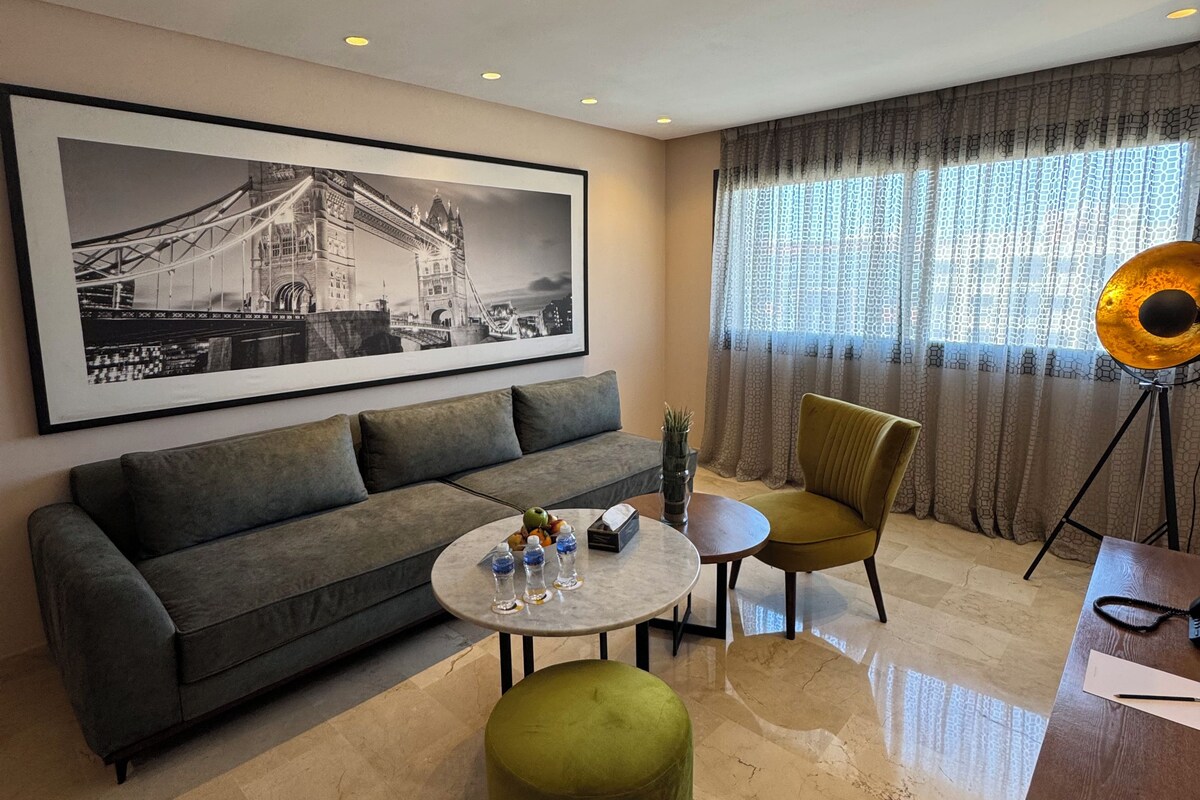 A modern living room with a sofa, a round coffee table, a green ottoman, a yellow armchair, and a tripod with a camera. A large framed picture of a bridge is on the wall.