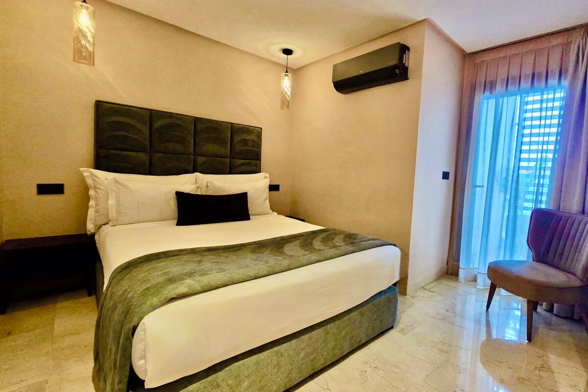 Habitación de hotel con una cama grande, cabecero, aire acondicionado de pared, ventana con cortinas y una silla.