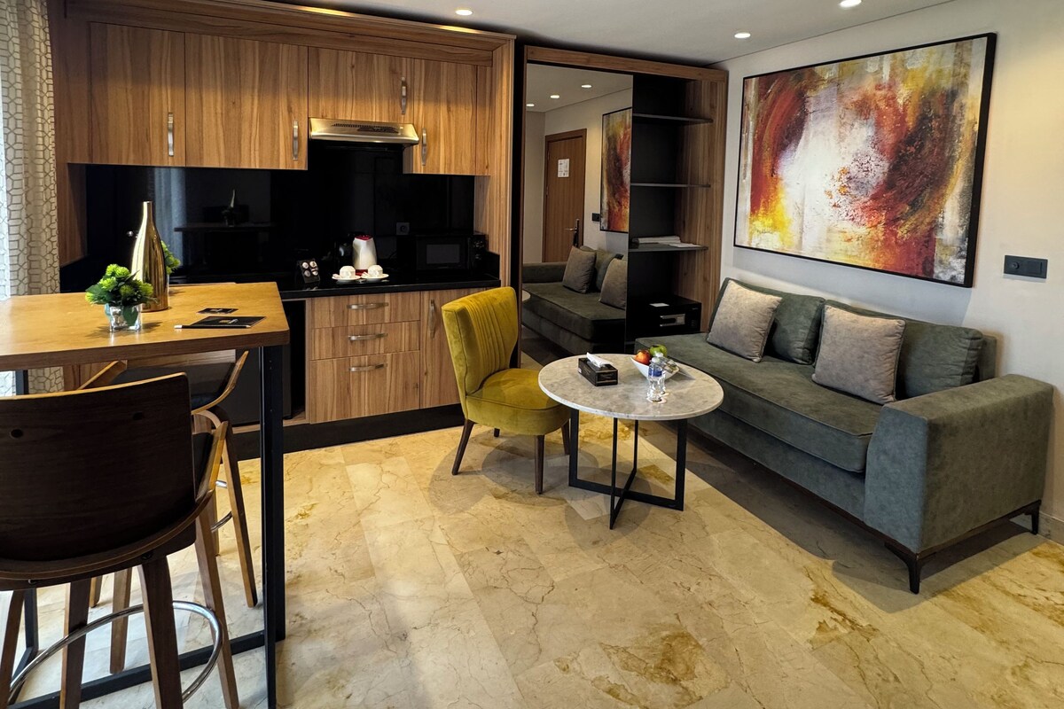 A modern living room with a grey sofa, a round coffee table, and a yellow armchair. There is a wooden dining table with chairs and a kitchen area with wooden cabinets.