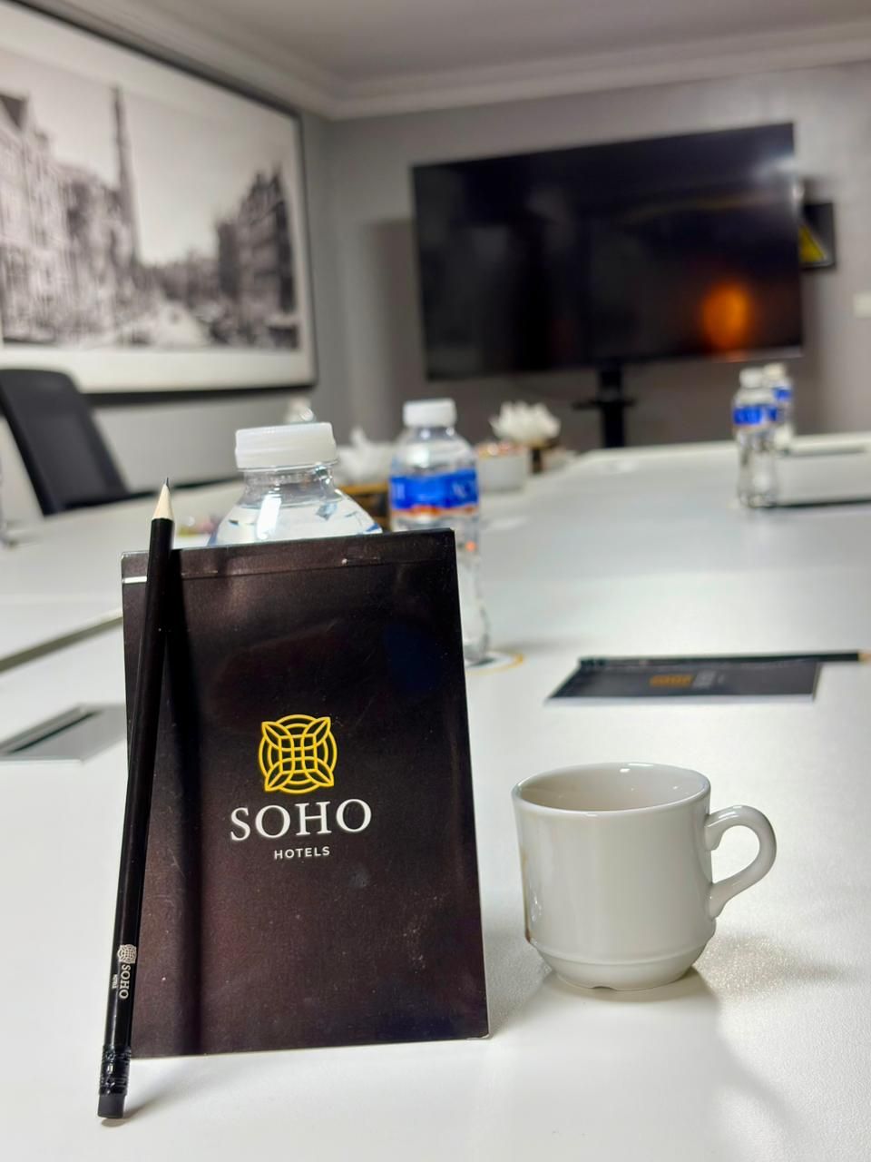 Quarto de hotel com uma mesa, um folheto preto da SOHO Hotels, uma caneca branca e garrafas de água.