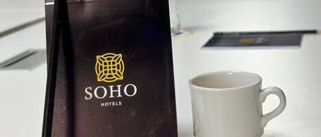 Quarto de hotel com uma mesa, um folheto preto da SOHO Hotels, uma caneca branca e garrafas de água.