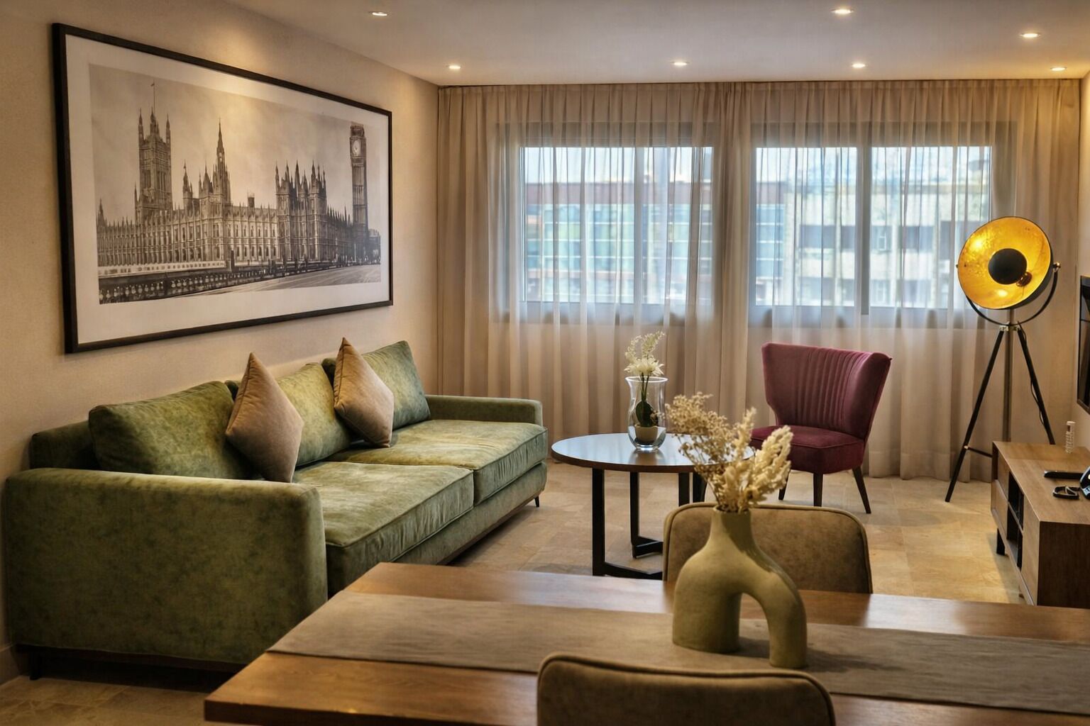 A modern hotel room with a green sofa, a red armchair, a round table, and a large window with curtains.