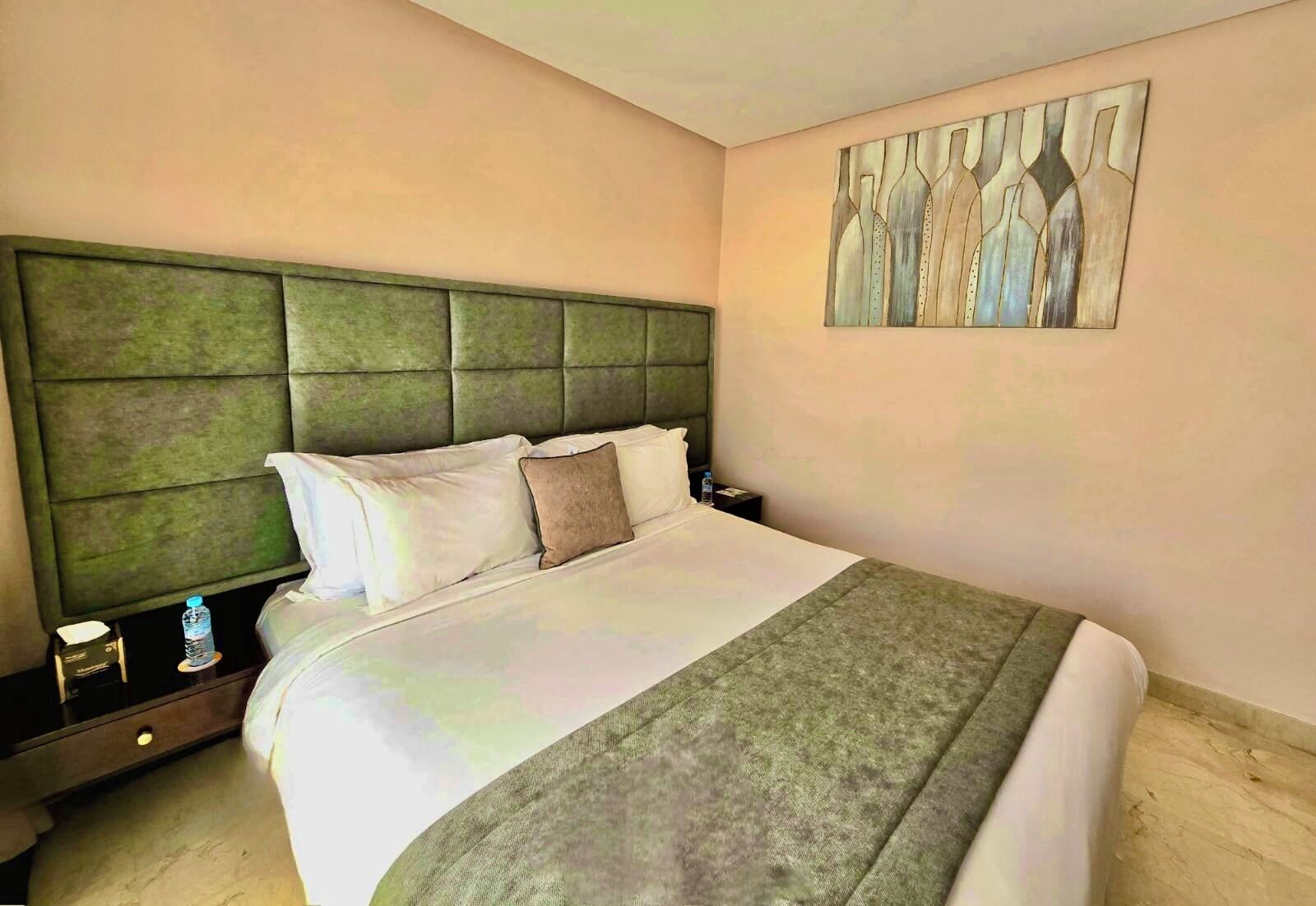 A hotel room with a large bed, a green headboard, a nightstand with a water bottle, and a painting on the wall.