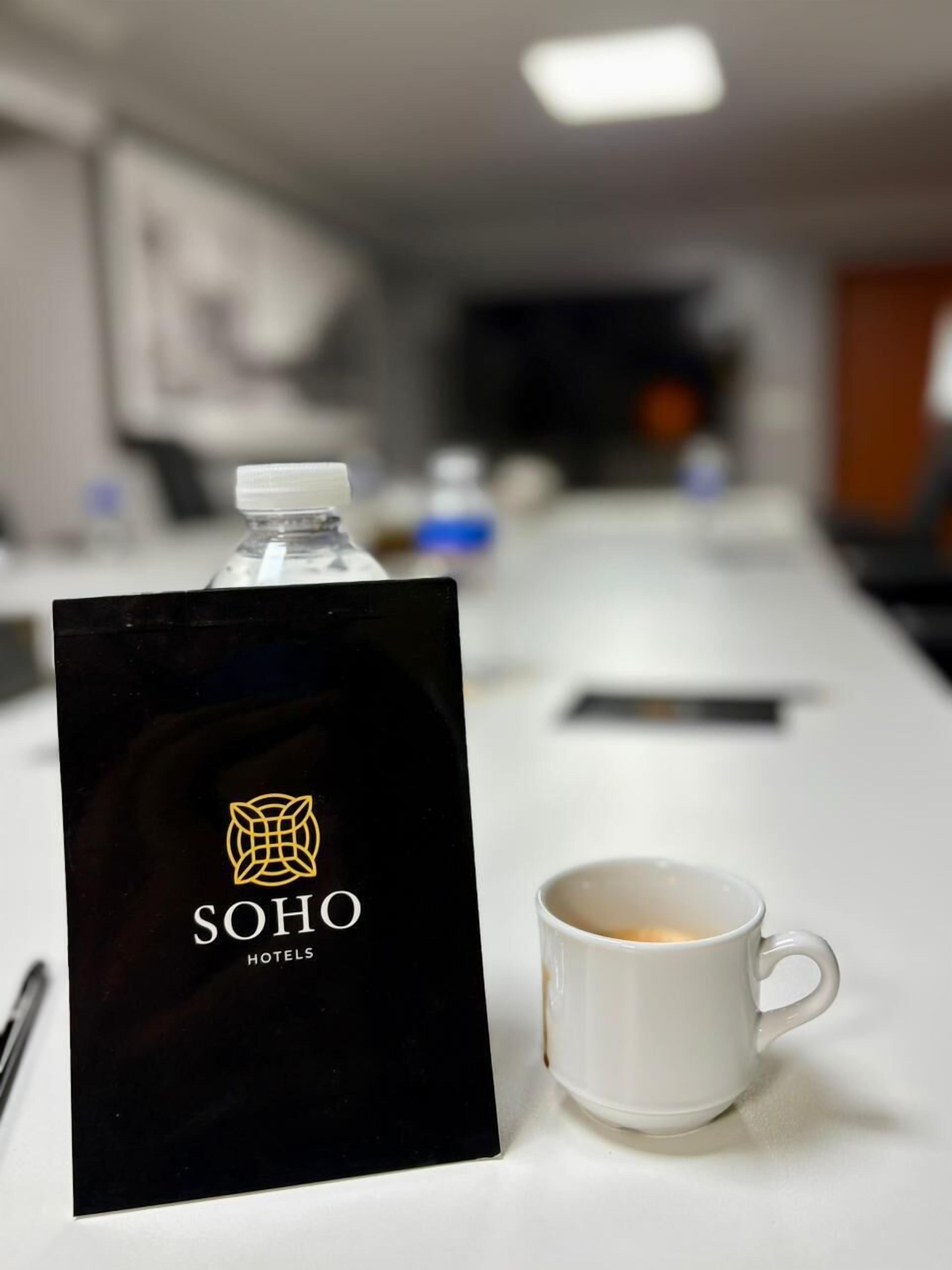 Ein Hotel-Empfangsbereich mit einem schwarzen Schild, das den Hotelnamen zeigt, einer weißen Tasse mit Kaffee und einer Wasserflasche auf einem Tresen.