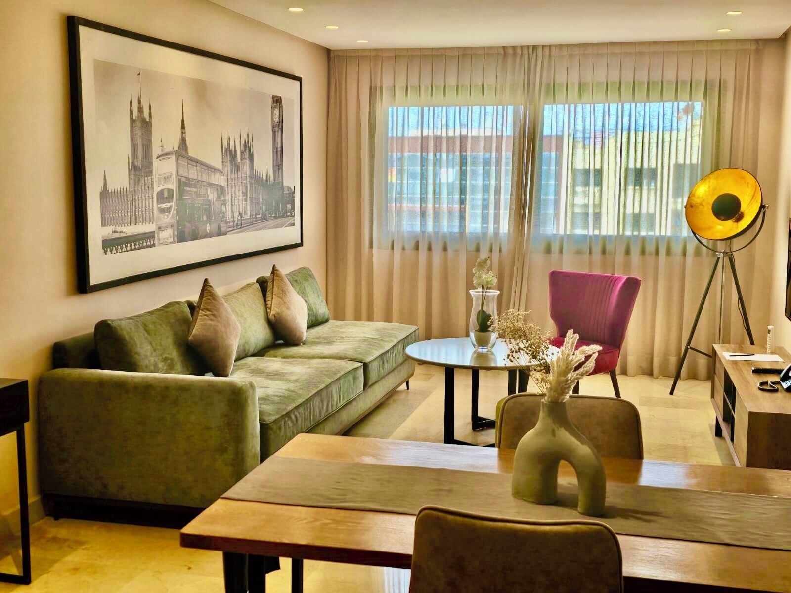 Een woonkamer met een bank, een tafel, een stoel, een vloerlamp, een ingelijste foto en een raam met gordijnen.