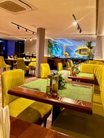Un restaurante con sillas y mesas amarillas, una zona de bar y una pared decorativa con palmeras.
