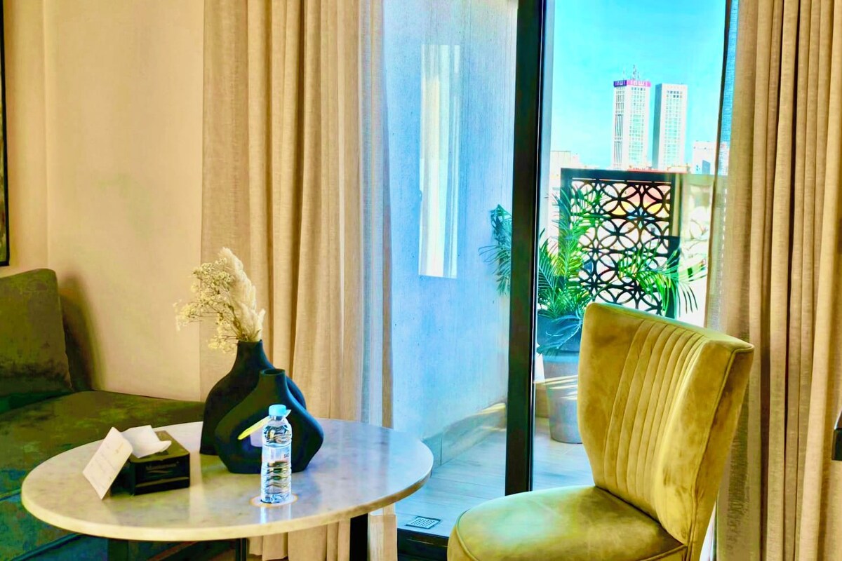 A modern hotel room with a round table, a sofa, a vase with flowers, and a view of tall buildings through a window with sheer curtains.