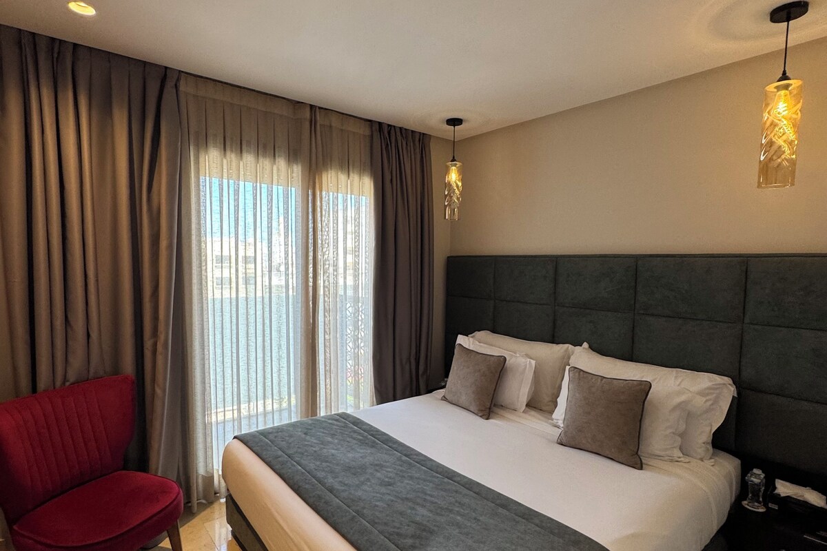 A hotel room with a large bed, a red chair, and a view through the window.